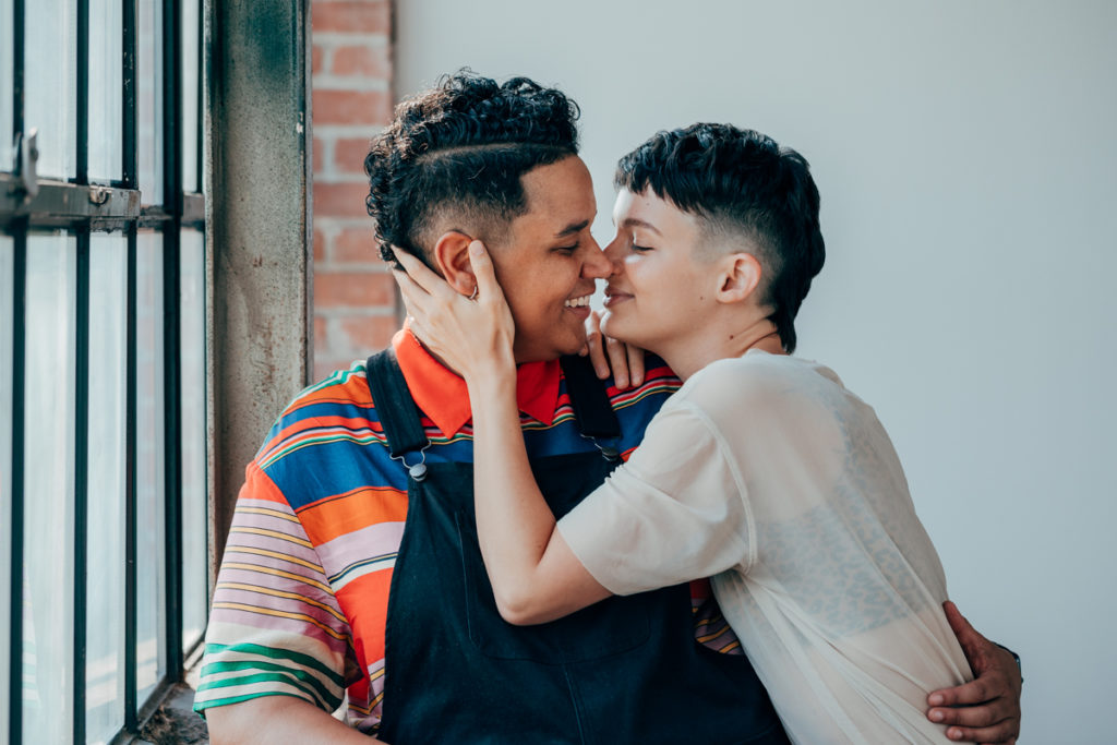 LGBTQ+ COUPLE DEEP ELLUM. They are in a loft with wonderful natural light. Both have fresh haircuts with fades. Emily is pulling heather in to kiss them. One is wearing overalls and the other is wearing a light colored t shirt with green pleated slacks.