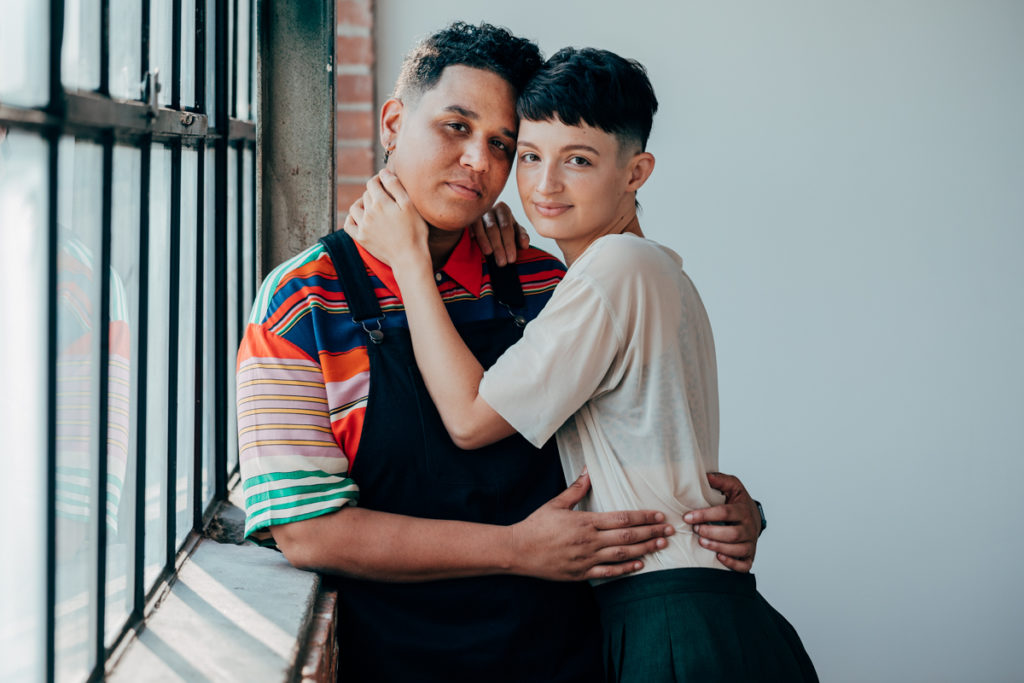 LGBTQ+ COUPLE DEEP ELLUM. They are in a loft with wonderful natural light. Both have fresh haircuts with fades. They are snuggled into each other and looking directly at the camera. One is wearing overalls and the other is wearing a light colored t shirt with green pleated slacks.
