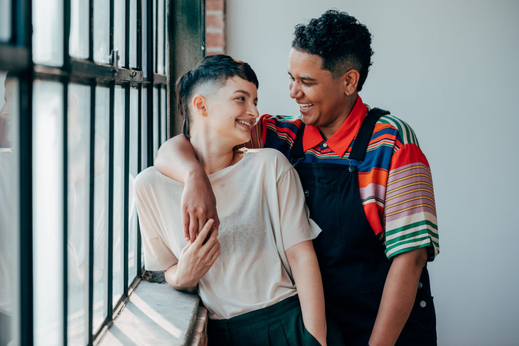 LGBTQ+ COUPLE DEEP ELLUM. They are in a loft with wonderful natural light. Both have fresh haircuts with fades. They are laughing at each other. One is wearing overalls and the other is wearing a light colored t shirt with green pleated slacks.