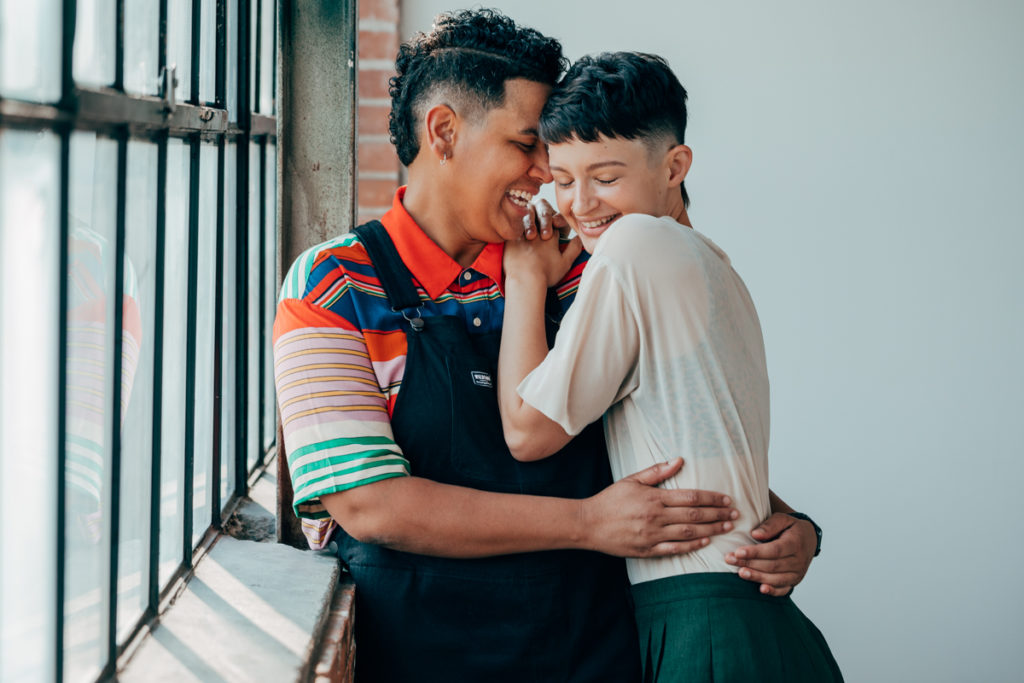 LGBTQ+ COUPLE DEEP ELLUM. They are in a loft with wonderful natural light. Both have fresh haircuts with fades. They are snuggled into each other. One is wearing overalls and the other is wearing a light colored t shirt with green pleated slacks.