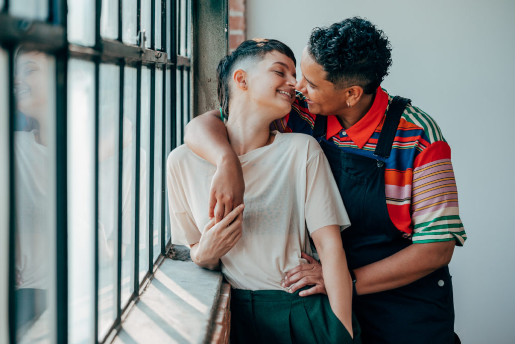 LGBTQ+ COUPLE DEEP ELLUM. They are in a loft with wonderful natural light. Both have fresh haircuts with fades. They facing each other and laughing about to kiss. One is wearing overalls and the other is wearing a light colored t shirt with green pleated slacks.