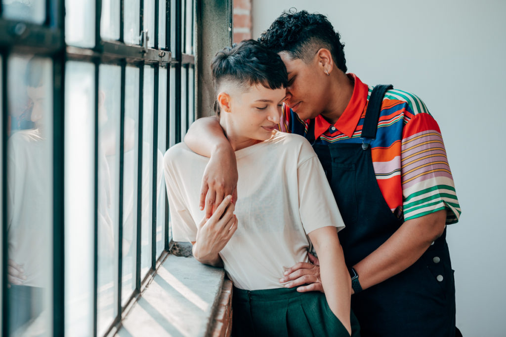 LGBTQ+ COUPLE DEEP ELLUM. They are in a loft with wonderful natural light. Both have fresh haircuts with fades. They are snuggled into each other. One is wearing overalls and the other is wearing a light colored t shirt with green pleated slacks.